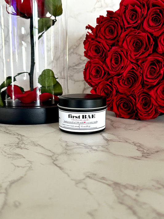 Black travel tin candle labeled “First BAE” on a marble surface beside a bunch of red roses and a single rose in a glass dome