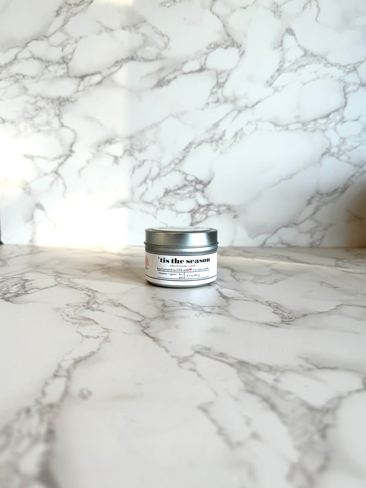 Silver travel tin candle labeled “tis the season” on a marble surface