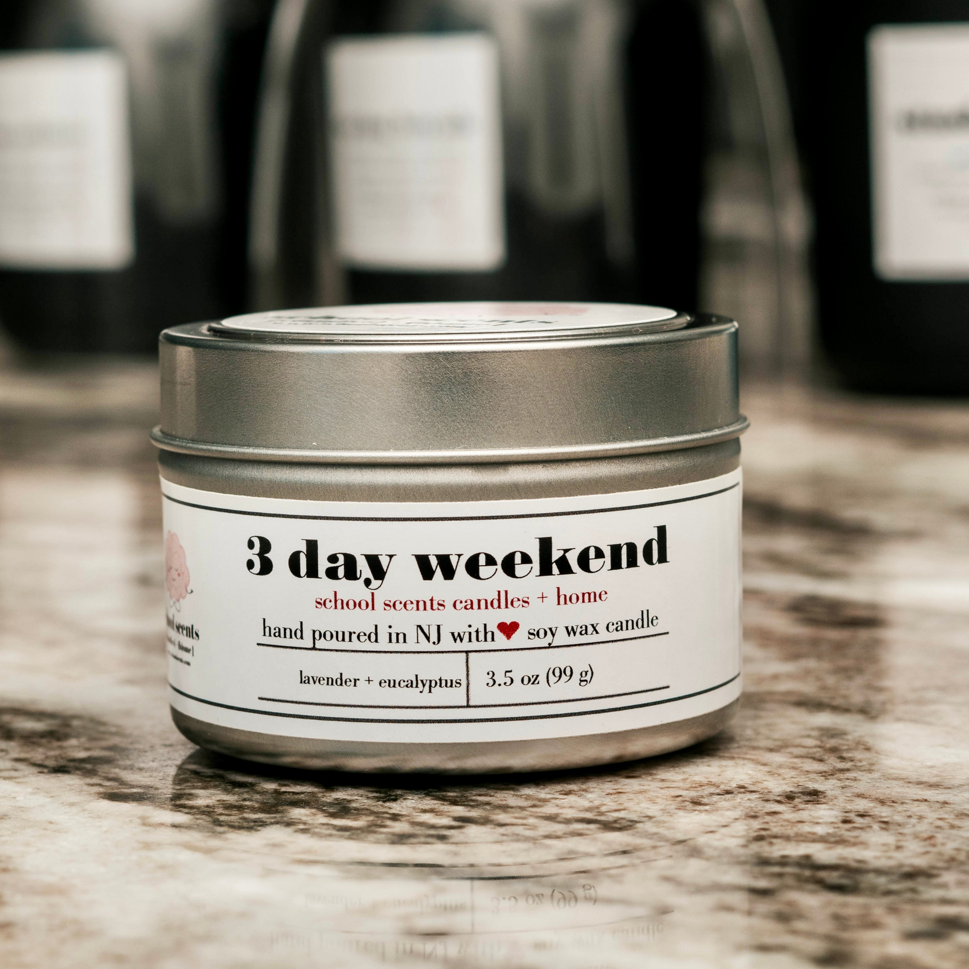 Close up of Silver travel tin candle labeled “3 day weekend” on a marble surface with blurred black matte candles in the background